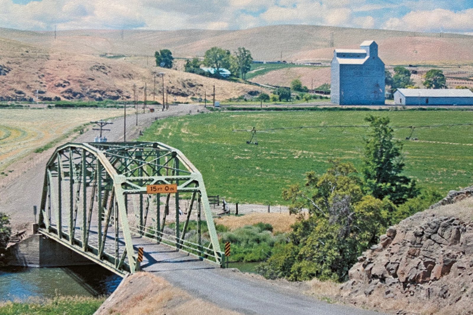 Steel truss bridge over the river, irrigated fields and a grain elevator beyond